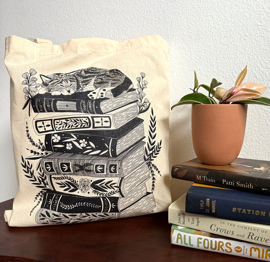 Tote bag with book design next to a stack of books and a potted plant.